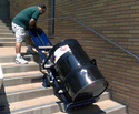 Man in green t-shirt and white shorts guiding a blue 2-wheeled powered dolly with a black 55 gal metal barrel on it up cement steps on the outside of a brick building